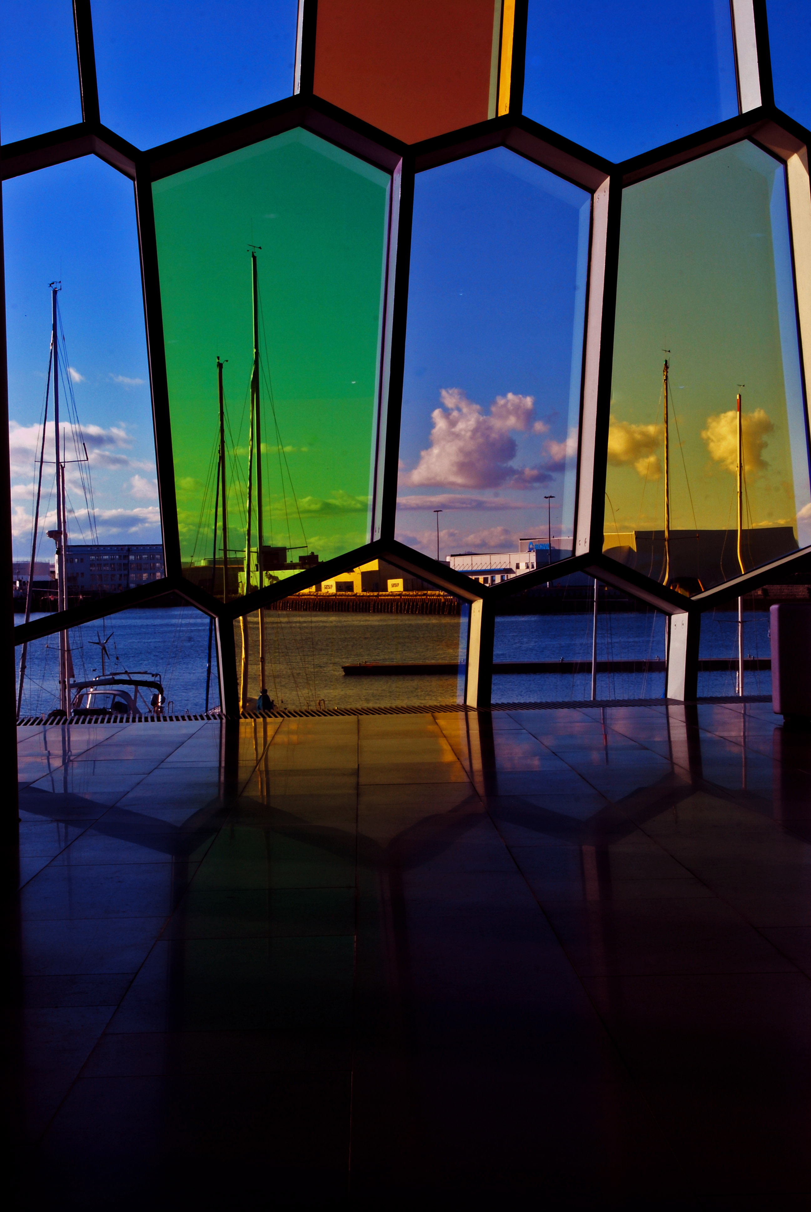 Vetrate colorate di forma irregolare che si riflettono sul pavimento lucido e dalle quali si vede il porto con le barche a vela, il mare e le nuvole